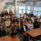 Group photo of about 30 students and instructors in a bright classroom around tables.