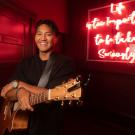 A man stands with an acoustic guitar and smiles at the camera. He appears to be backstage at a club, with a neon saying over his shoulder that read 'Life is too important to be taken seriously.'