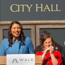 A woman stands at a podium in front of city hall while people listen.