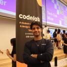 A student in a black long sleeve T-shirt, stands in front of a black banner that reads “code lab”, in a lecture hall filled with other students. 