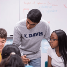 Student wearing UC Davis shirt leans over to speak to younger students in classroom setting