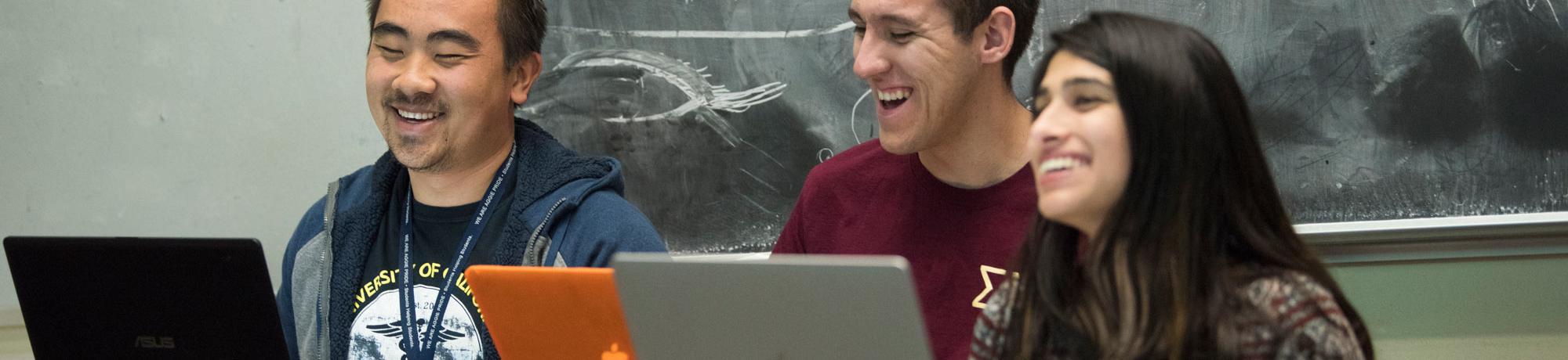 three students sitting in front of a chalkboard with laptops laughing in a classroom