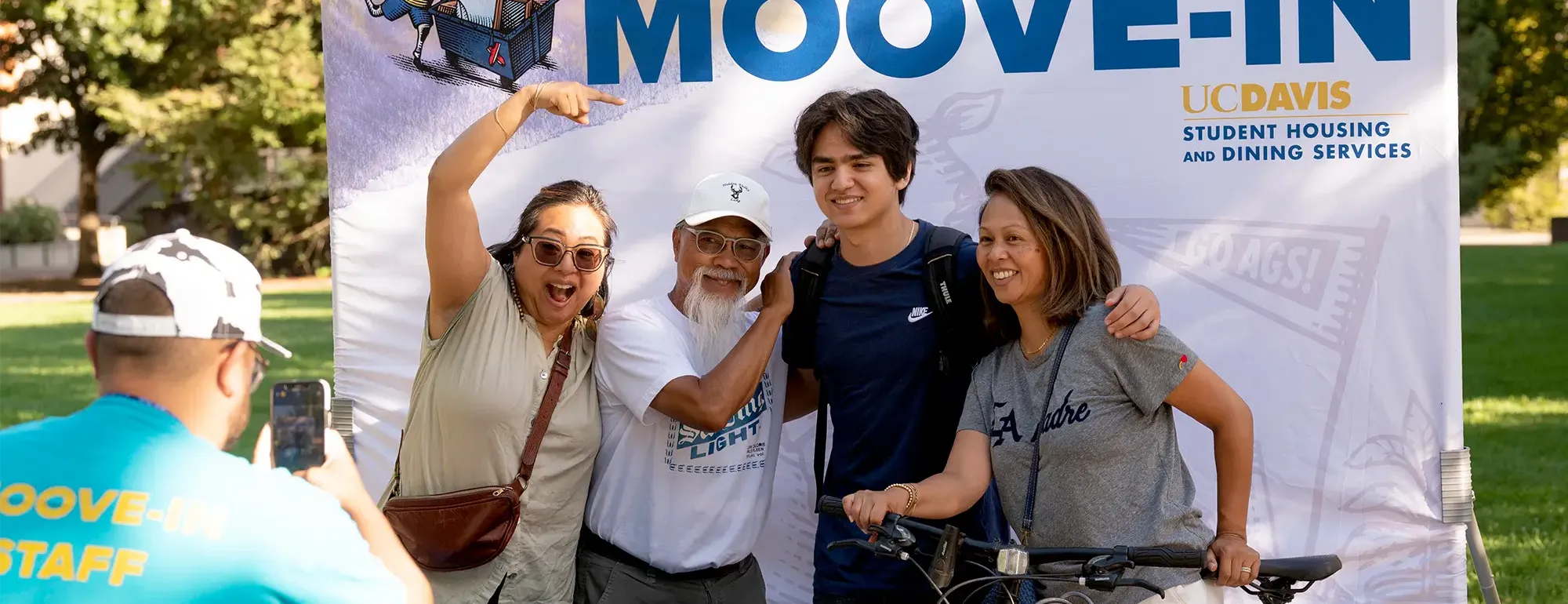 Family posing for a photo with a student in front of a 'Welcome to Moove-In' UC Davis sign, while a staff member takes their picture outdoors.